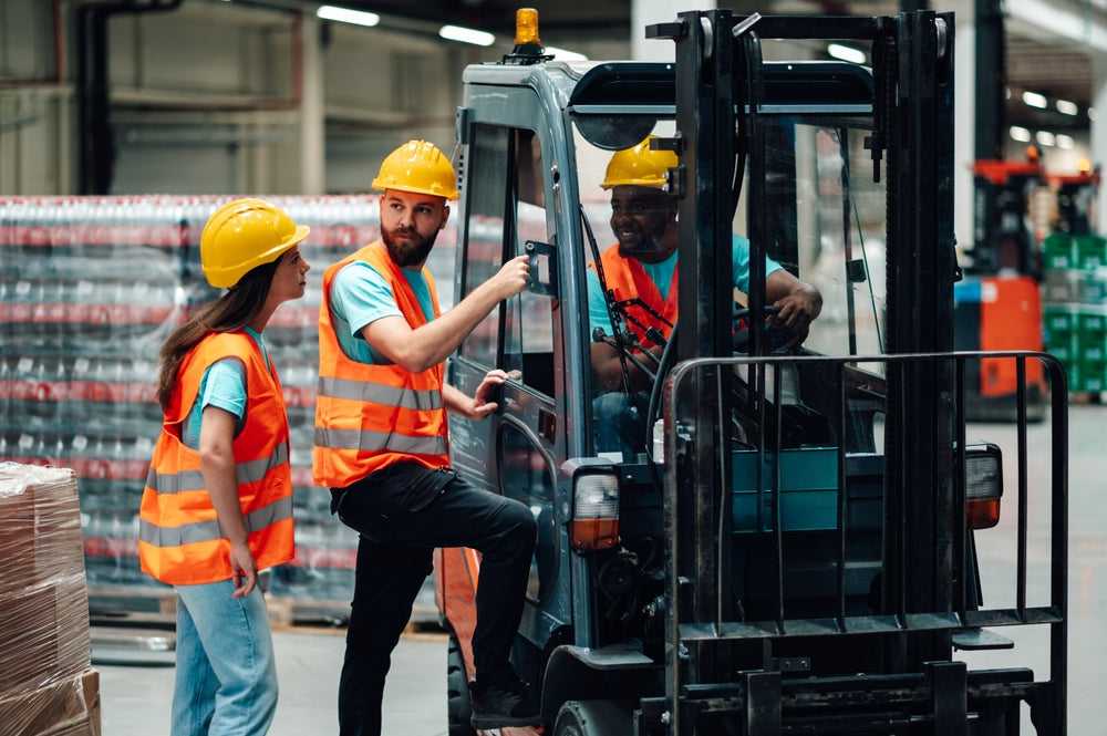 Warehouse workers wearing safety vests and hard hats are discussing forklift safety training in a bottling plant