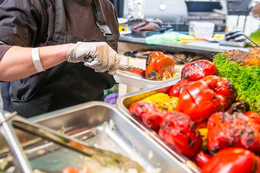 Food service worker wearing gloves uses tongs to plate up brightly colored peppers
