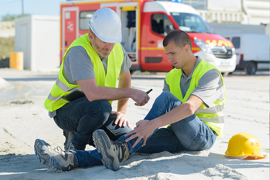 Two workers are kneeling down looking at one workers leg, there is an ambulance in the background